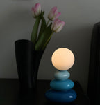 Blue Clérachic table lamp lit in a dim room, with a black vase of pink tulips beside it.