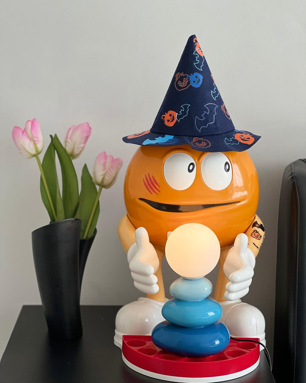 A blue-pebbled Clérachic table lamp glowing in front of a large orange character figure in a wizard hat, with a vase of tulips beside.