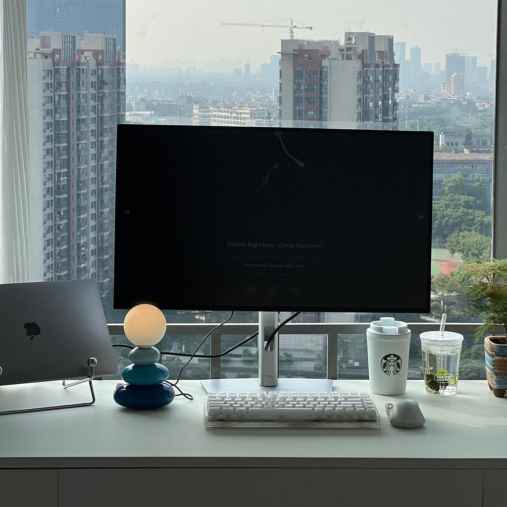 Small blue Clérachic table lamp on a white desk below a monitor, with a laptop, keyboard and drinks cups; a city skyline shows through the window.