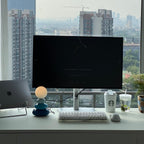Small blue Clérachic table lamp on a white desk below a monitor, with a laptop, keyboard and drinks cups; a city skyline shows through the window.