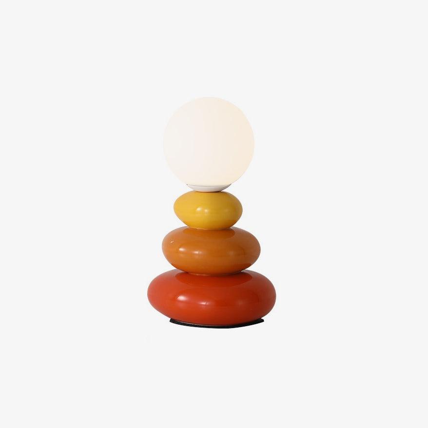 Cut-out style view of this Clérachic table lamp, its pale globe over three pebbles in yellow, orange and red, on a plain off-white ground.