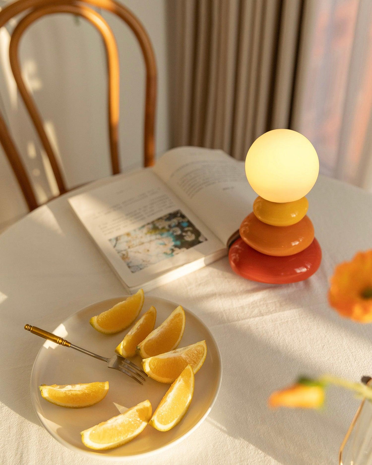 Top-lit Clérachic table lamp on a clothed table, with an open book, a plate of orange slices and a bentwood chair in warm light.