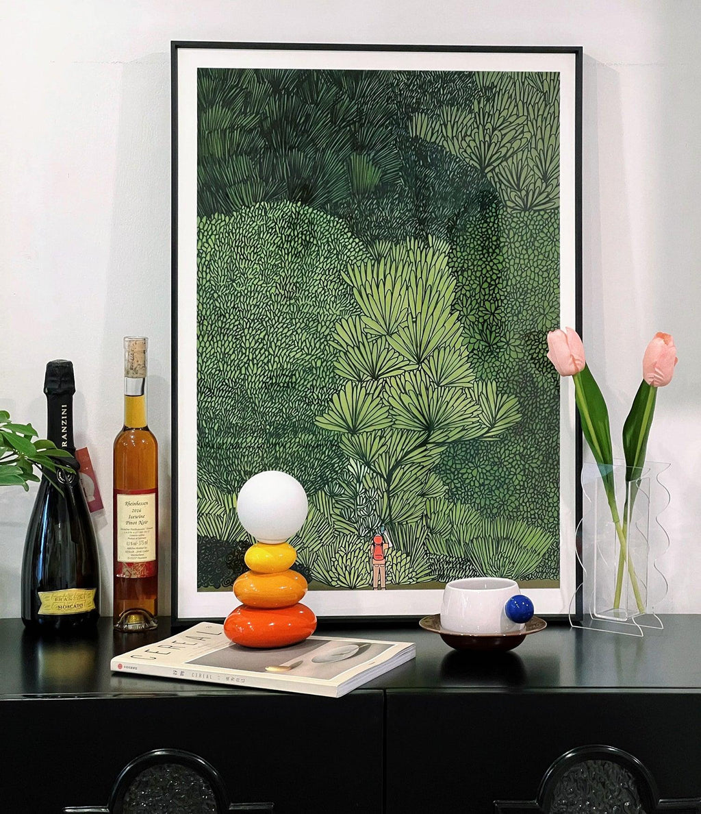 Small Clérachic table lamp on a black sideboard in front of a large leafy print, with bottles, tulips and a cup beside it.