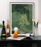 Small Clérachic table lamp on a black sideboard in front of a large leafy print, with bottles, tulips and a cup beside it.