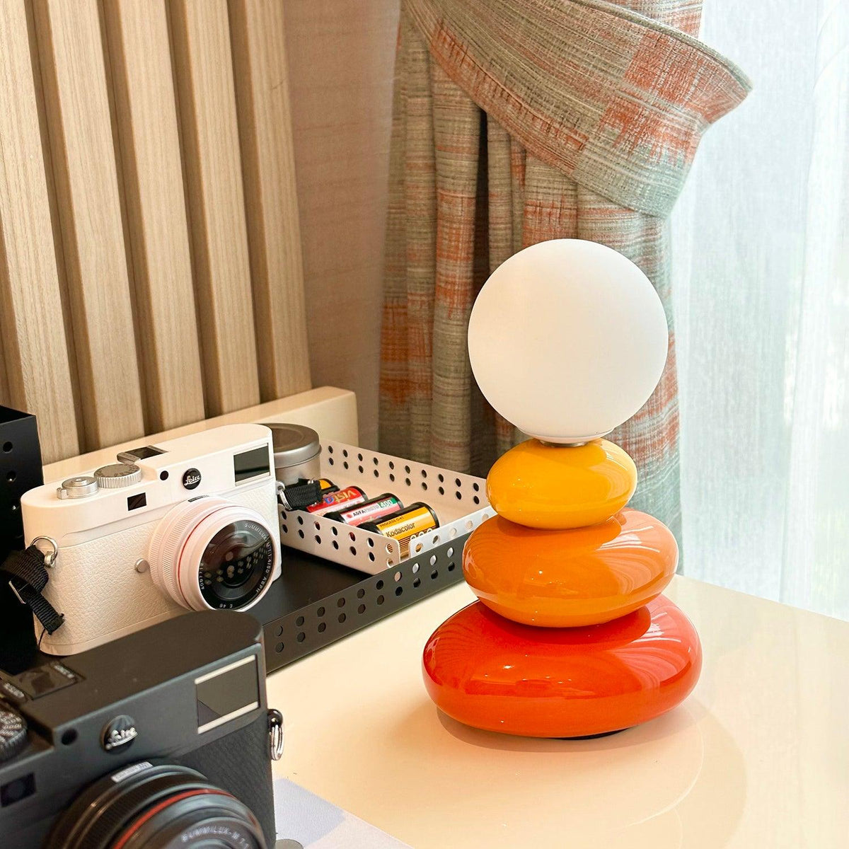 This Clérachic table lamp on a cream desk in front of a striped curtain, with a white camera and a tray of batteries behind.