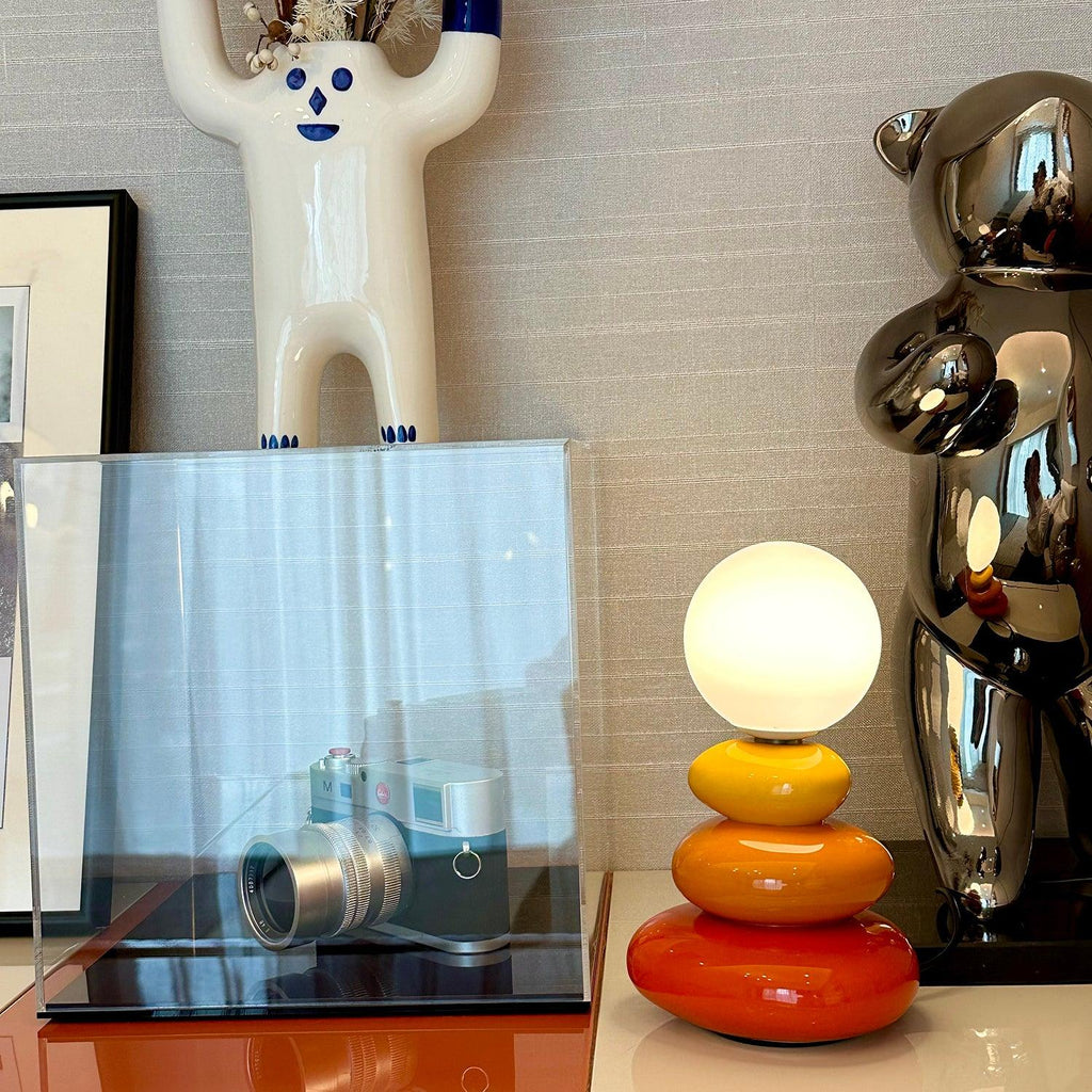 Orange and red Clérachic table lamp lit on a shelf, next to a clear display box holding a silver camera and a chrome bear figure.