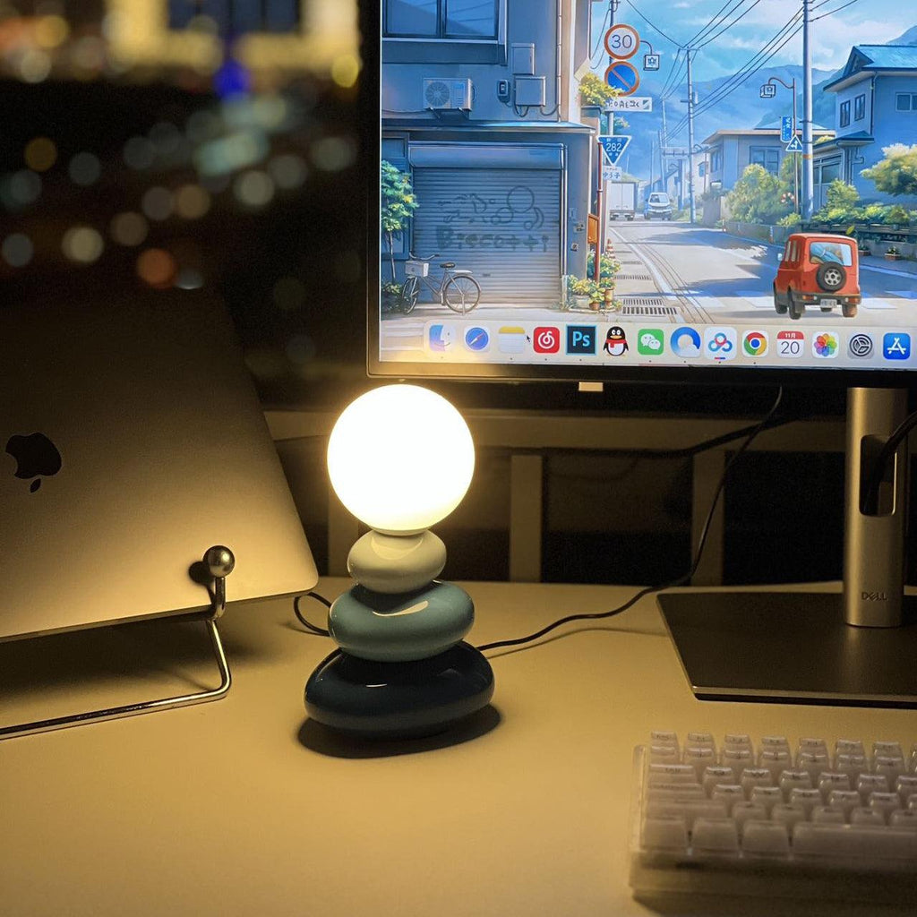 Dark teal-grey Clérachic table lamp lit warm on a desk, with a monitor showing a street scene, a laptop and a keyboard.