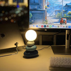 Dark teal-grey Clérachic table lamp lit warm on a desk, with a monitor showing a street scene, a laptop and a keyboard.
