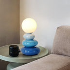 Blue Clérachic table lamp, lit, on a pale green round side table with a small black speaker, beside a grey sofa arm.