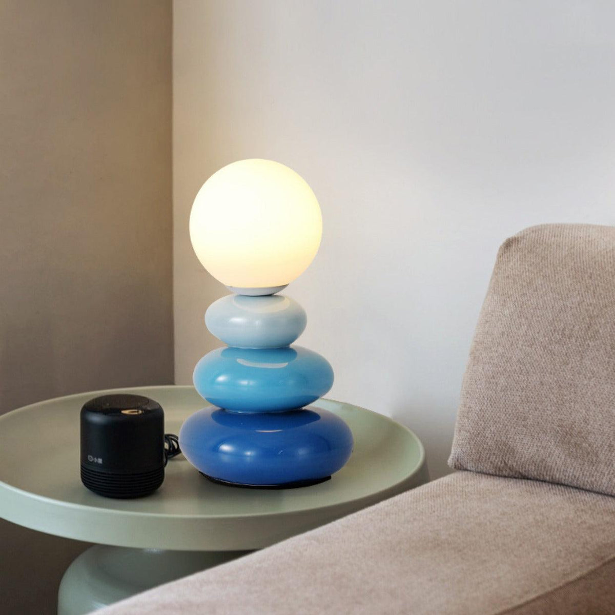 Blue Clérachic table lamp, lit, on a pale green round side table with a small black speaker, beside a grey sofa arm.