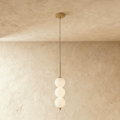 Clérachic three-ball pendant in brushed gold with opal glass spheres, lit and hanging against a limewash wall.