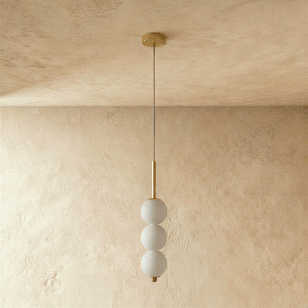 Clérachic gold pendant with three frosted glass spheres on a slim stem, switched off against a warm plaster wall.
