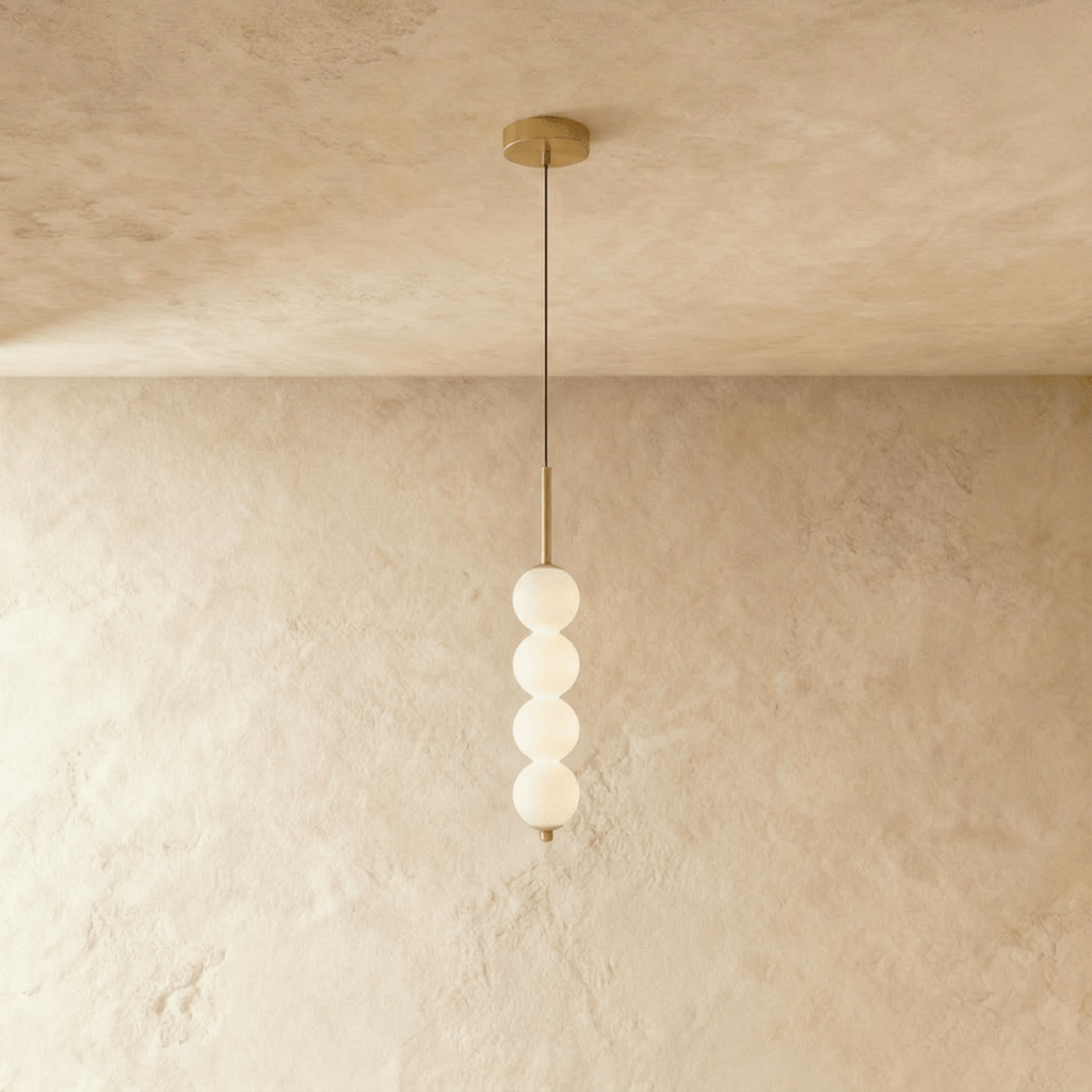 Clérachic four-ball pendant in gold with glowing opal glass spheres hanging from a thin cord on a plaster wall.