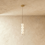 Clérachic four-ball pendant in gold with glowing opal glass spheres hanging from a thin cord on a plaster wall.