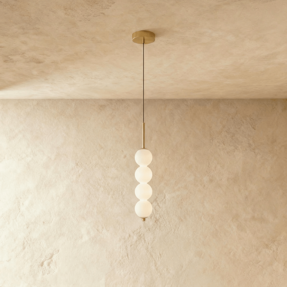 Clérachic four-ball pendant in gold with glowing opal glass spheres hanging from a thin cord on a plaster wall.