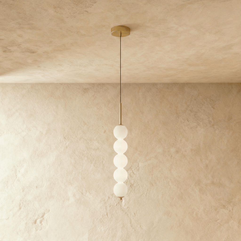 Clérachic five-ball pendant in gold with a stack of glowing opal glass spheres on a long cord, lit against plaster.