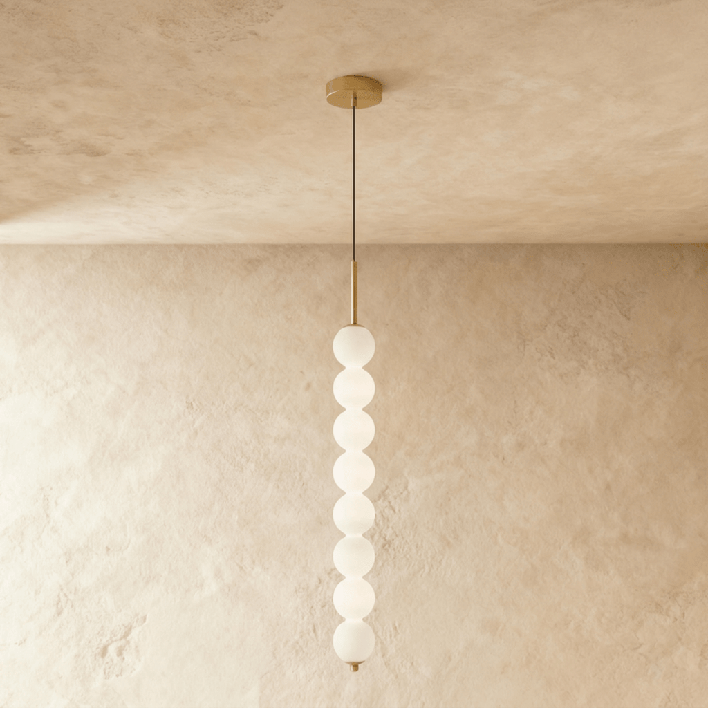 Clérachic eight-ball pendant in gold, a tall column of lit opal glass spheres hanging against a limewash wall.