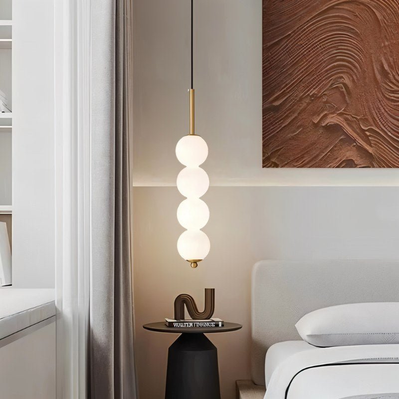 Clérachic gold four-ball pendant glowing beside a bed, with a round side table, stacked books and a textured art panel.