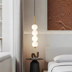 Clérachic gold four-ball pendant glowing beside a bed, with a round side table, stacked books and a textured art panel.