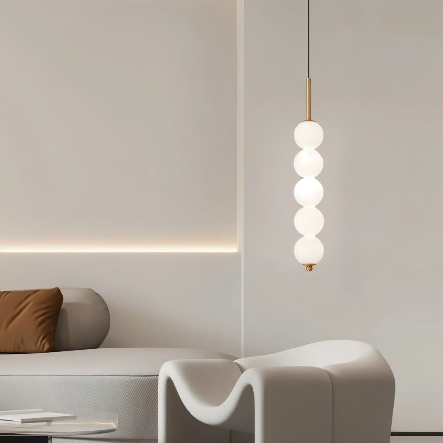 Clérachic gold five-ball pendant glowing in a pale living room with a sculptural white armchair and a cove-lit wall.