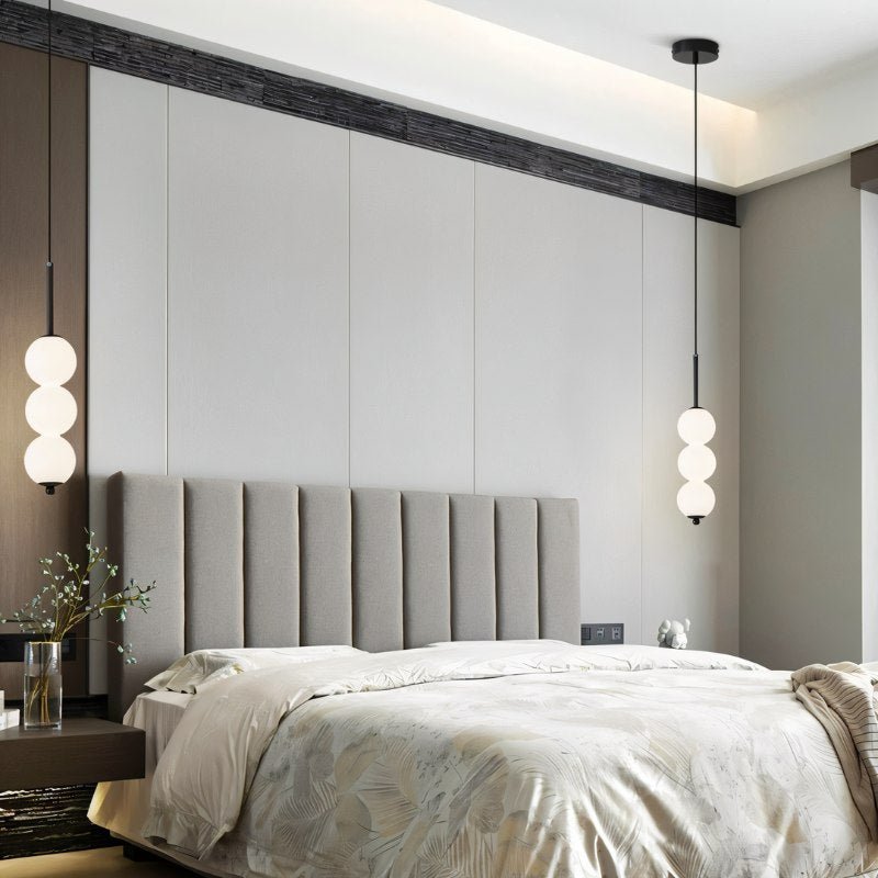 Two Clérachic black three-ball pendants flanking a grey upholstered headboard in a calm bedroom with a panelled wall.