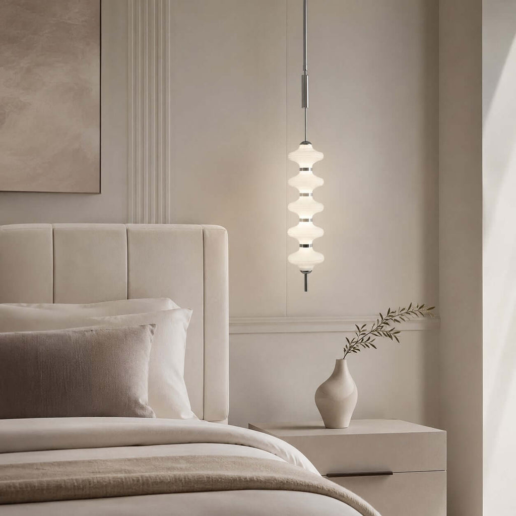 Chrome and white Clérachic pendant of stacked ribbed discs glowing beside an upholstered cream headboard and a bud vase.