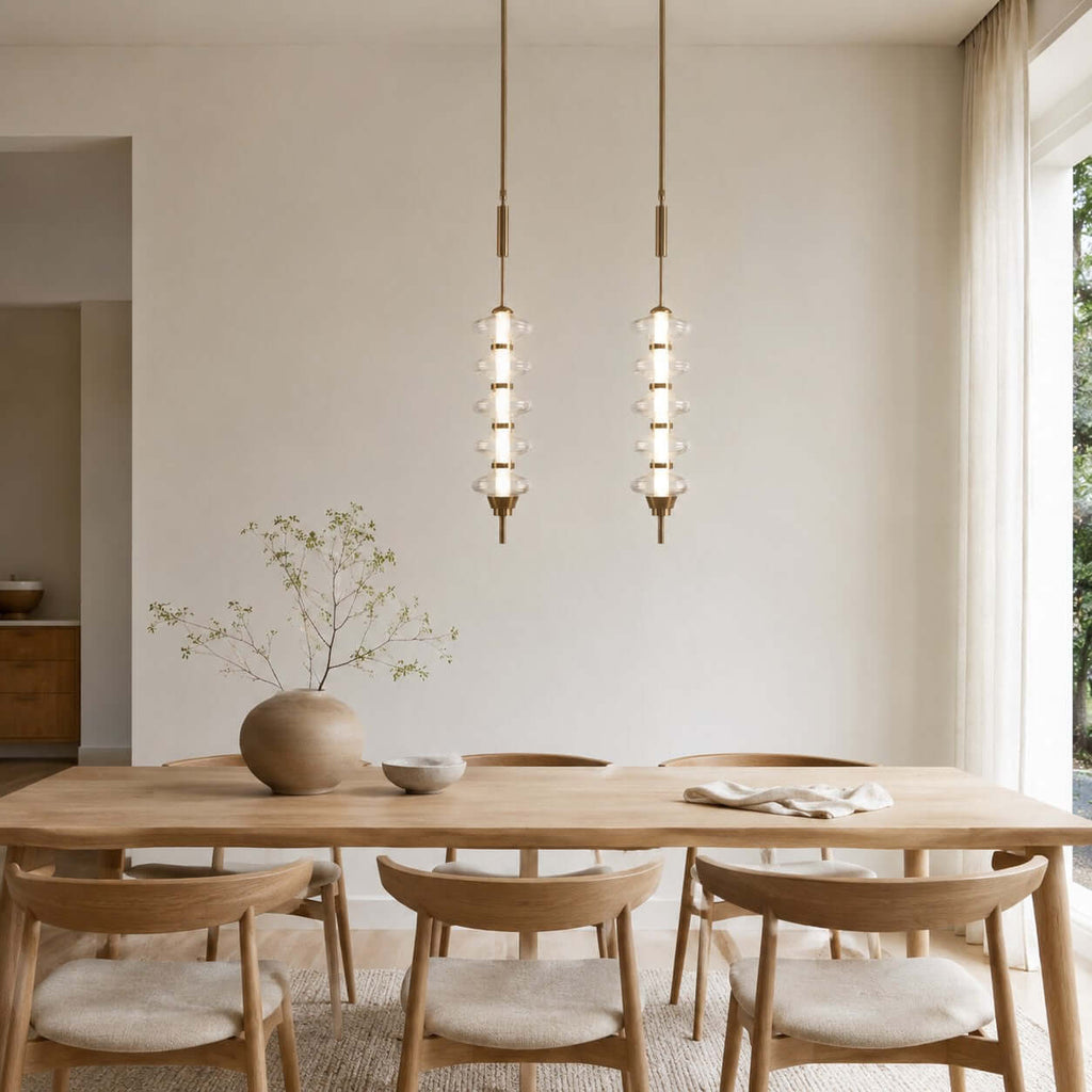 Two gold and clear Clérachic pendants, tall and slim with stacked glass discs, hung above a pale oak dining table.