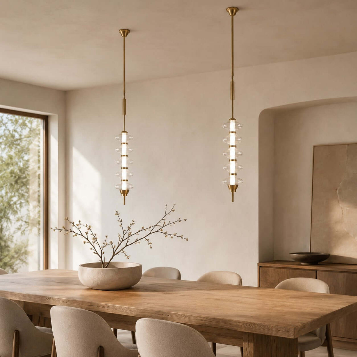 A pair of gold and clear Clérachic pendants of stacked glass discs, hanging in a plastered dining room with a window and arched niche.