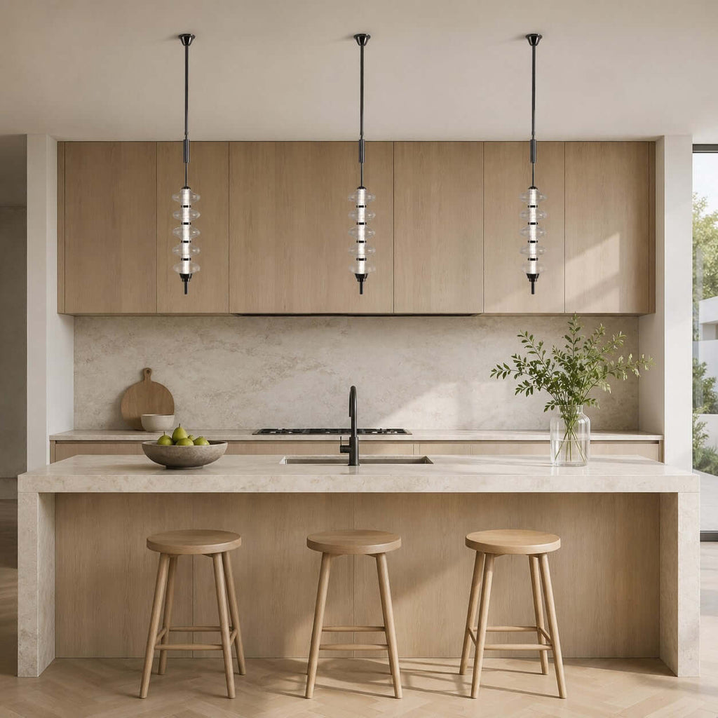 Three black and clear Clérachic pendants in a row over a stone kitchen island with wooden stools, against oak cabinets.