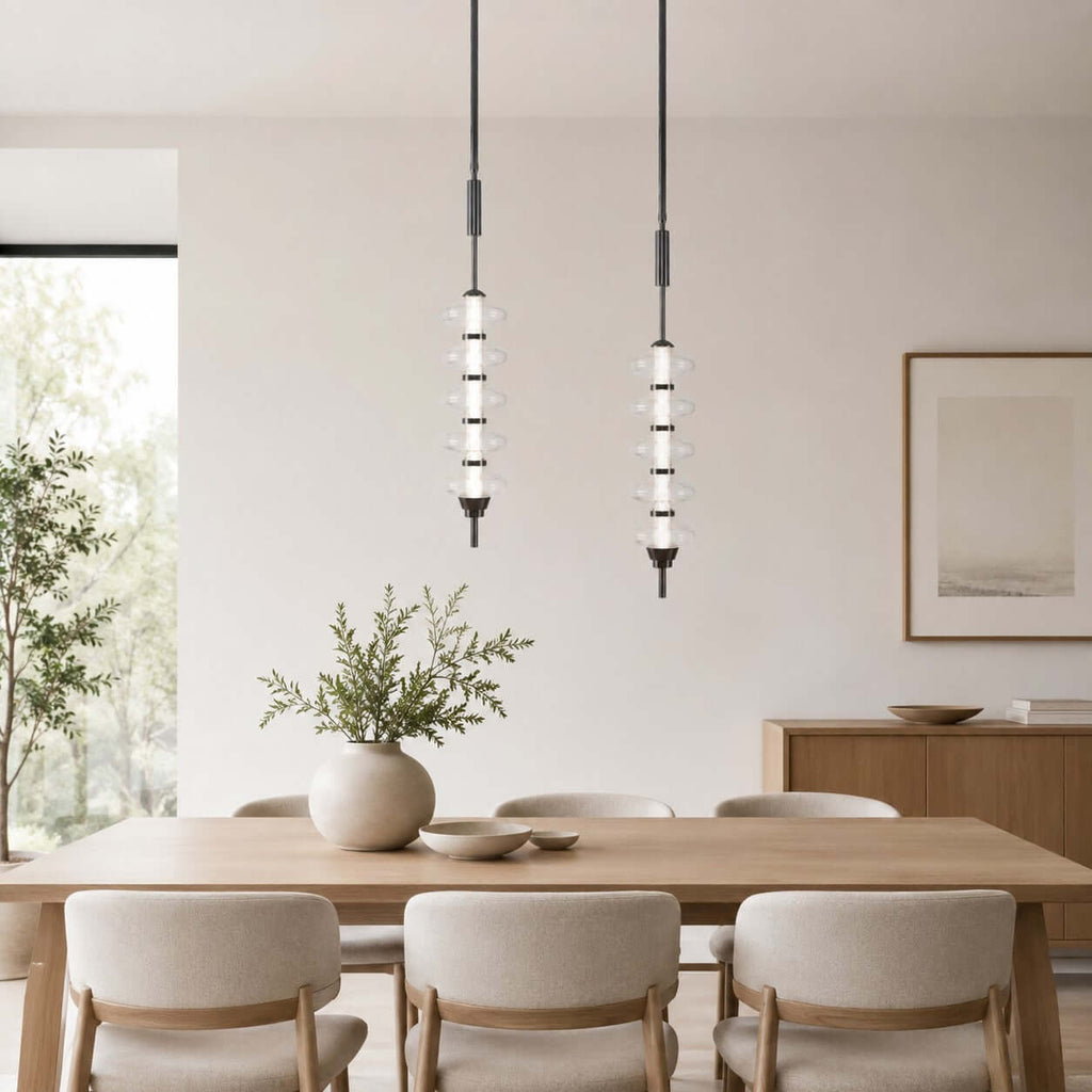 Two black and clear Clérachic pendants over a light wooden dining table, a vase of greenery and cream chairs below.