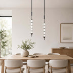 Two black and clear Clérachic pendants over a light wooden dining table, a vase of greenery and cream chairs below.