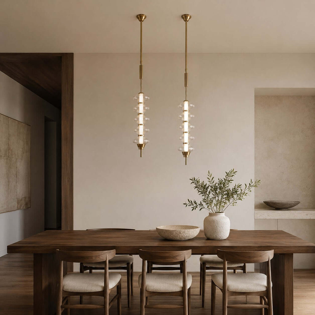 Gold and clear Clérachic pendants side by side over a dark wood dining table, with a stone bowl and a vase of olive branches.