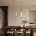 Gold and clear Clérachic pendants side by side over a dark wood dining table, with a stone bowl and a vase of olive branches.