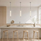 Three chrome and white Clérachic pendants in a row over a stone island with four pale wood stools, a window to one side.