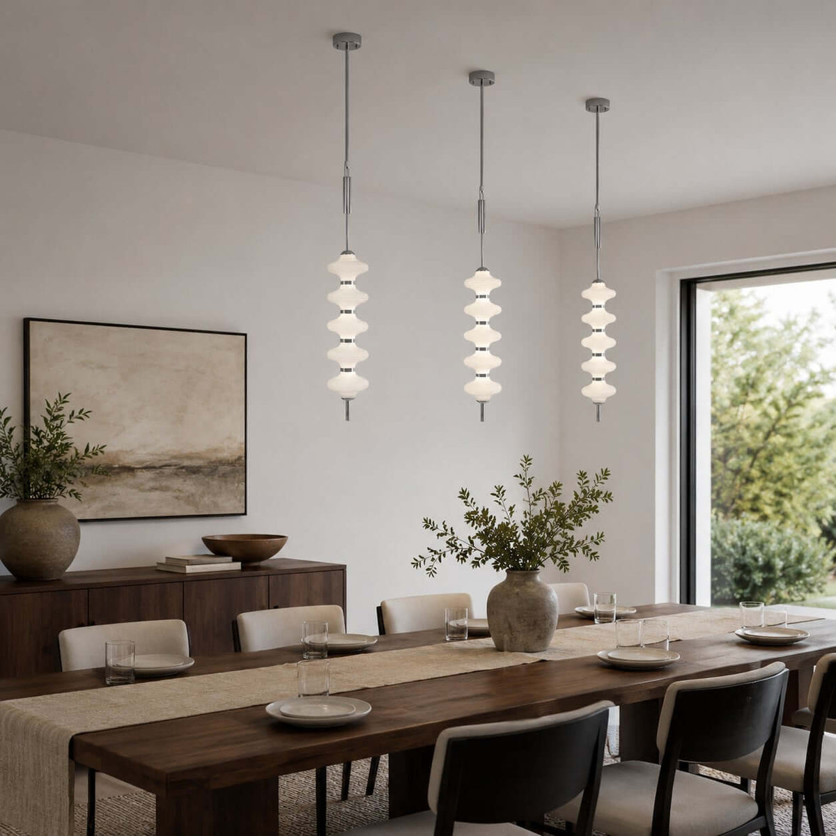 Three chrome and white Clérachic pendants at different heights above a long dark dining table, beside a framed landscape.