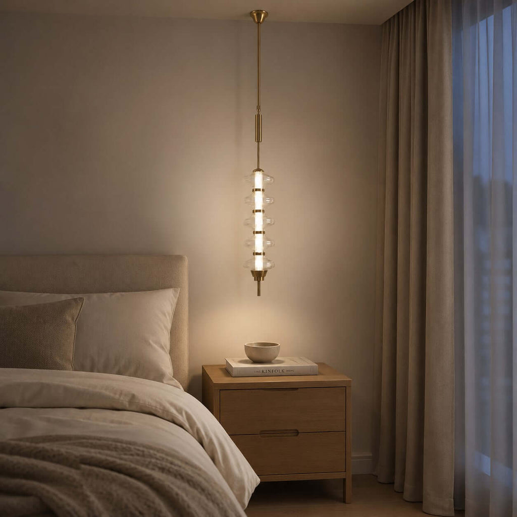 Gold and clear Clérachic pendant glowing at bedside level next to a wooden nightstand, with heavy curtains and a rumpled bed.