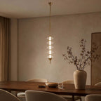 Gold and clear Clérachic pendant lit at dusk over a dark wood dining table, with a tall vase of dried branches beside it.