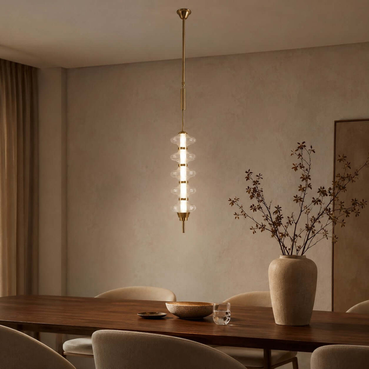 Gold and clear Clérachic pendant lit at dusk over a dark wood dining table, with a tall vase of dried branches beside it.