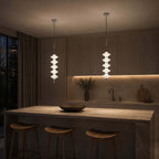 Chrome and white Clérachic pendants lit in a dim kitchen at night, over a stone island with wooden stools.