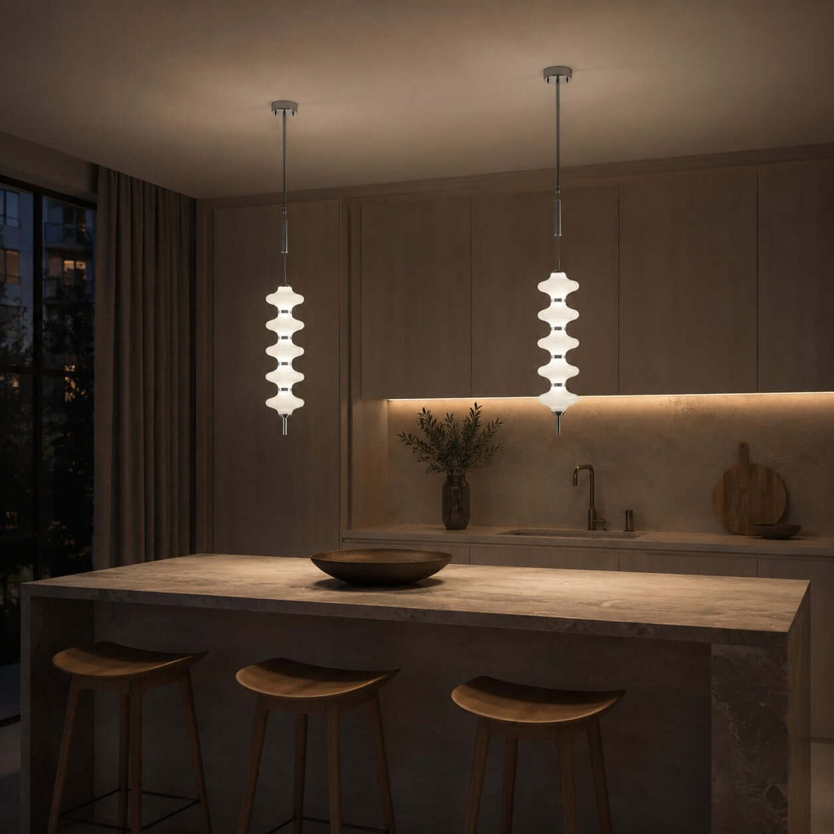Chrome and white Clérachic pendants lit in a dim kitchen at night, over a stone island with wooden stools.