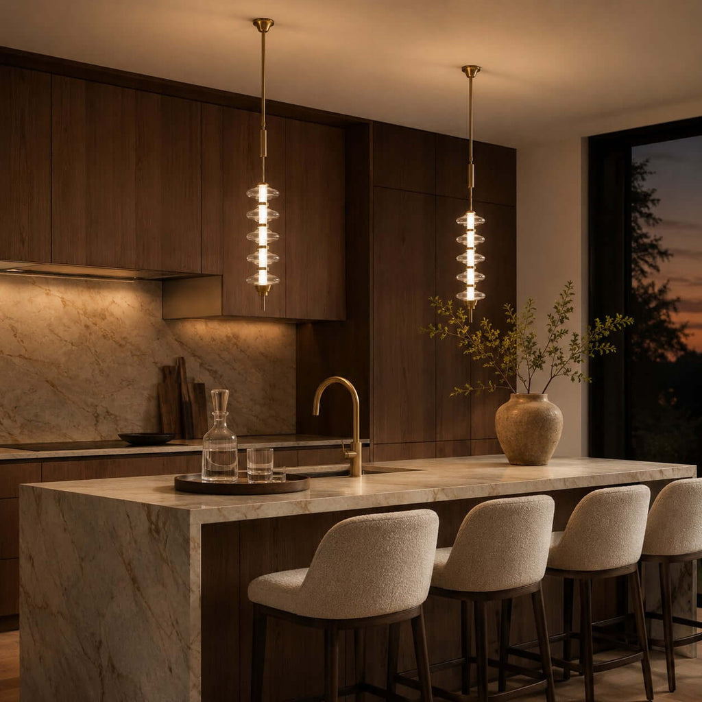 Two gold and clear Clérachic pendants glowing over a marble island with boucle bar stools, a sunset behind the window.