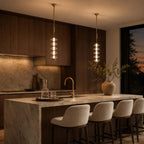 Two gold and clear Clérachic pendants glowing over a marble island with boucle bar stools, a sunset behind the window.