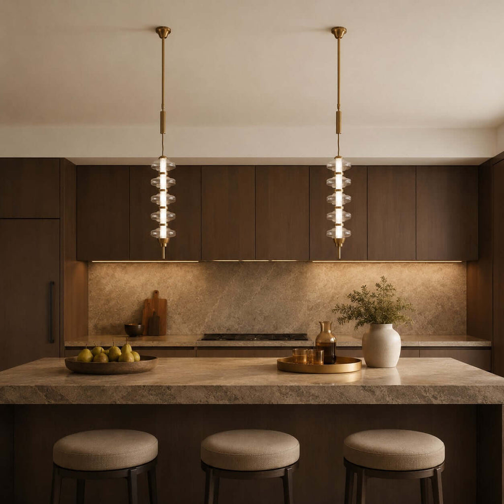 A pair of gold and clear Clérachic pendants lit above a stone island and bar stools, with a fruit bowl on the counter.