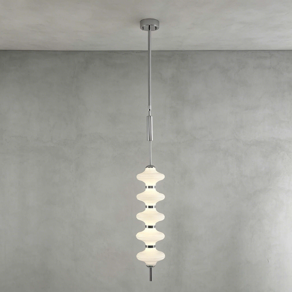 Chrome and white Clérachic pendant of stacked ribbed frosted discs, lit and hanging from its canopy against a grey plaster wall.