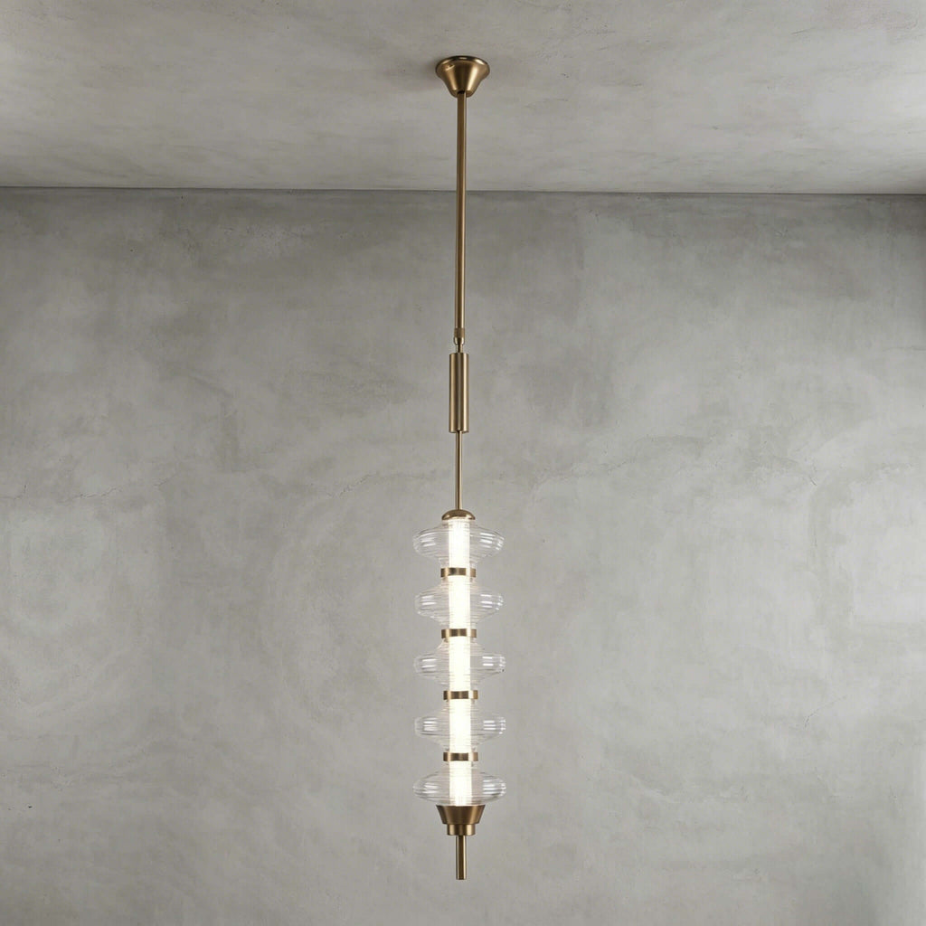 Gold and clear Clérachic pendant with stacked clear discs around a lit centre, on a long gold rod against a grey plaster wall.