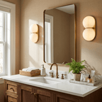 A pair of Clérachic glass sconces glowing warm cream either side of a brass-framed mirror above a walnut vanity with a fern.