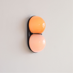 Angled view of a black-backed Clérachic wall light with an orange upper globe and a peach lower globe on a pale wall.