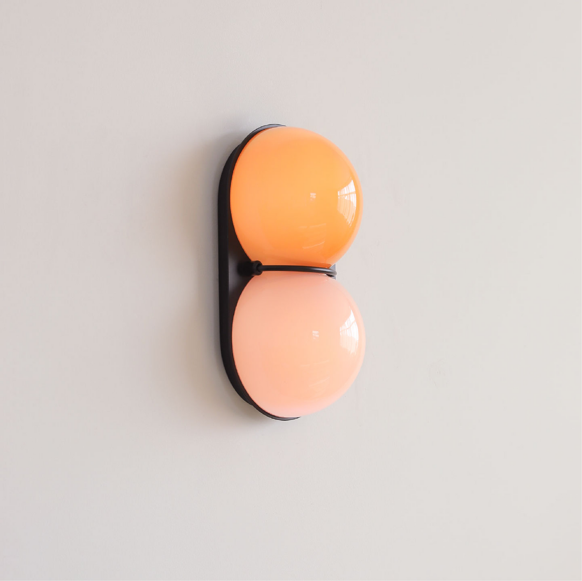Angled view of a black-backed Clérachic wall light with an orange upper globe and a peach lower globe on a pale wall.