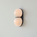 Lit from within, the two globes of this Clérachic wall light glow a soft peach against a black oval plate and pale wall.