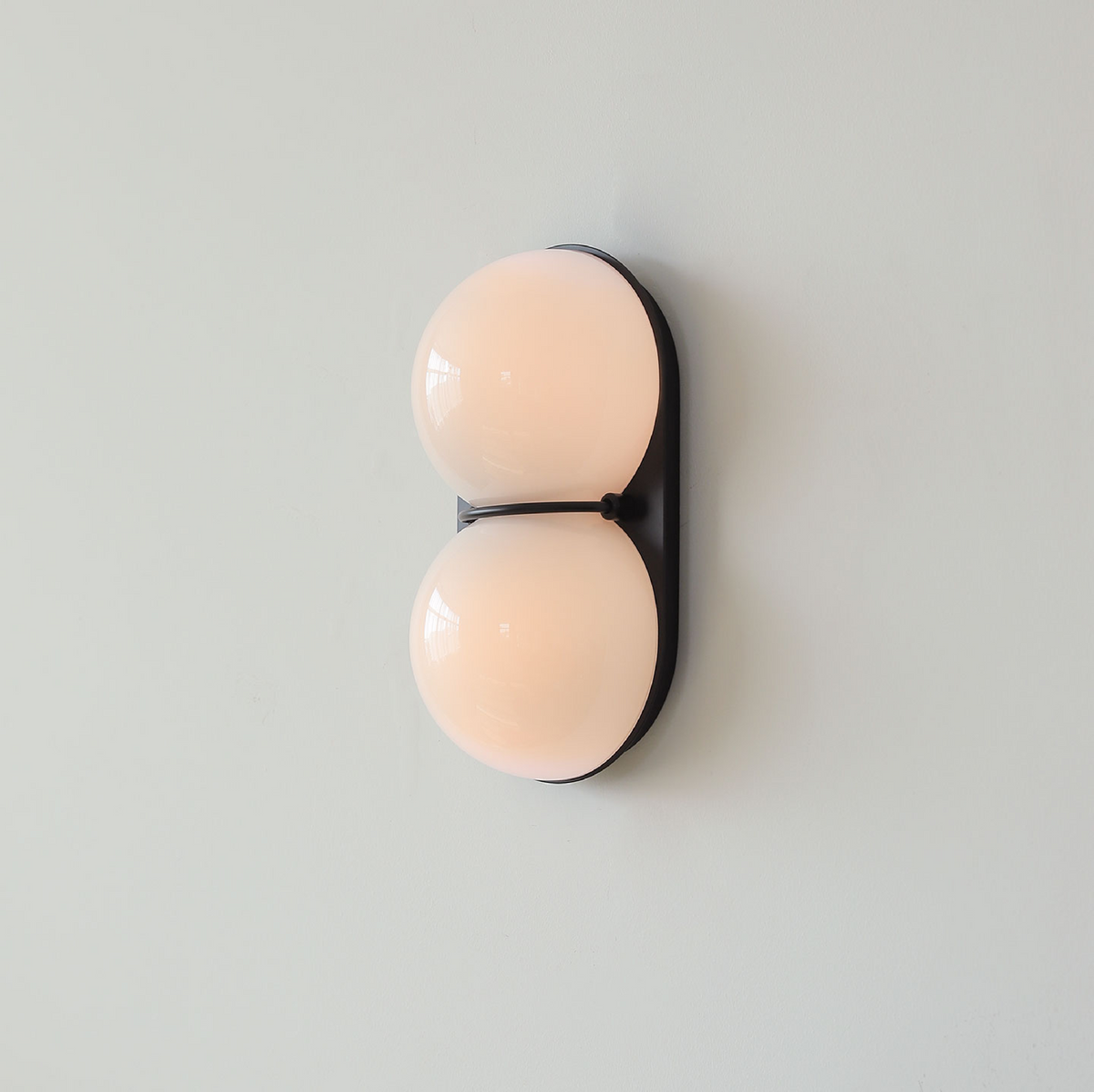 Lit from within, the two globes of this Clérachic wall light glow a soft peach against a black oval plate and pale wall.